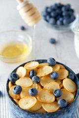 Blue Bowl of mini pancake cereal with blueberries, maple syrup and milk on concrete background. Tiny pancakes, new food trend concept. Cute healthy breakfast or snack. Recipe, menu.