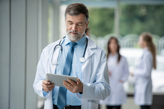 Confident smiling doctor standing with a tablet and looking at camera in a hospital. Medical team standing behind doctor