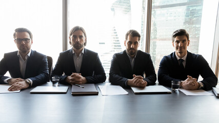 Group of focused male professional headhunters recruiters sitting at table, waiting for job candidate for interview. Serious hr managers teammates in formal wear looking at camera, hiring process. - obrazy, fototapety, plakaty