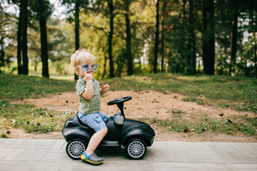 Little pretty caucasian boy with short fair hair in summer clothes rides a toy black car in the big park