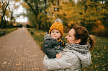 Young beautiful mother in warm clothes plays with her young Caucasian son in the autumn park