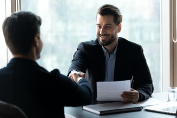 Smiling young handsome businessman in formal wear shaking hands with male confident partner after...