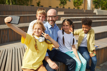 Group of cheerful teenage kids taking selfie with smiling male teacher outdoors in sunlight, copy space
