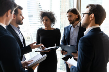 Focused young multiracial diverse business people in formal wear standing in circle, holding clipboard with paper reports, sharing project ideas opinions at informal conversation in modern office.
