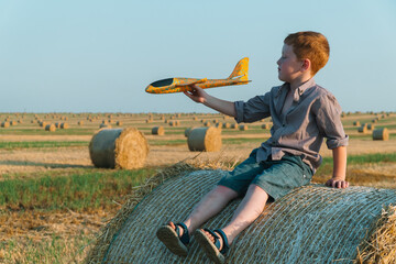Naklejka na ściany i meble The red-haired boy plays with a toy airplane on a wheat field with bales