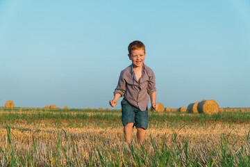 Naklejka na ściany i meble The red-haired boy goes ahead of a wheat field on a background of straw bales