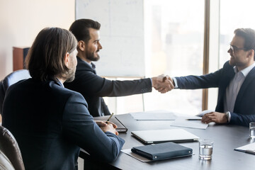 Happy businessman watching two confident male partners shaking hands after discussing agreement contract details at boardroom table, making cooperation decision, celebrating successful cooperation.