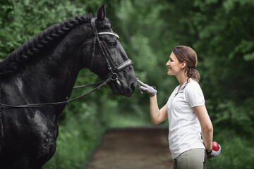 portrait of beautiful young woman standing near black friesian stallion in forest