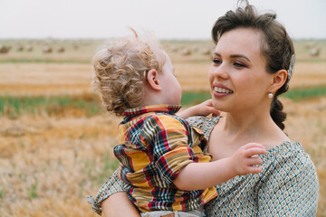 Fototapeta na wymiar Beautiful young woman with fair-haired little son in her arms in the middle of a wheat field with bales in summer evening