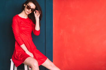Beautiful fashion girl with long hair, spanish appearance in sunglasses and red elegant dress posing on blue red wall in studio. Amazing babe in suglasses with black hair. Hairstyle advertsing.