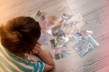 The boy lies on the floor in the apartment and looks at his printed on paper photos. The child wants to insert a photo into a frame and hang it on the wall.