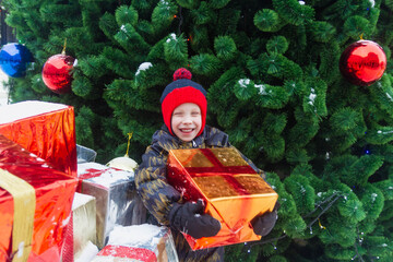 A European boy holds a large box with a Christmas gift and laughs in front of a Christmas tree decorated with ball.