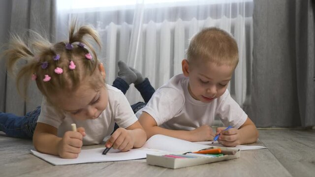 Cute children draw with crayons on white paper lying on the floor in the room.
