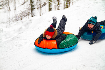 Fototapeta na wymiar Two European boys roll down a slide on snowtubes in the winter in the forest. Children's games in the snow. Snowy winter in Russia.