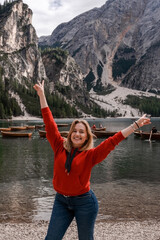 Dolomites Alps. Lago di Braies. Italy. Young blonde woman in red sweater & blue jeans feels freedom on background of  boats on the water, rock mountains & wooded foothills