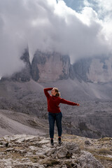 Dolomites Alps. Italy. Young blonde woman in red sweater & blue jeans feels freedom on background of Tre Cime di Lavaredo mountains with peaks wrapped by grey clouds