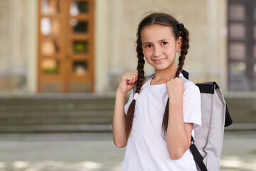 Waist up portrait of cute schoolgirl carrying backpack and smiling at camera while posing outdoors against school building, copy space