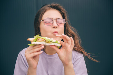 Young stylish trendy woman isolated over grey blue background. Girl enjoying eating tasty delicious sandwich during lunch or breakfast. Portrait of meal in hands.