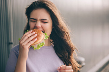Young stylish trendy woman isolated over grey blue background. Hungry girl biting piece of burger. Lunchtime or dinner. Tasty delicious burger in hand.