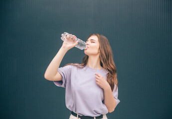Young stylish trendy woman isolated over grey blue background. Girl drinking clear tasty water from transparent plastic bottle. Body hydration.