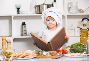 Smiling girl is looking new recipe in big book in the kitchen at home.