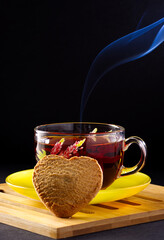 Glass cup with black tea on a saucer, with heart-shaped cookie. A smoke comes from the cup.