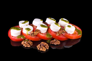 Tasty salad with tomato, goat cheese, pesto and walnuts  with reflection, isolated on black background