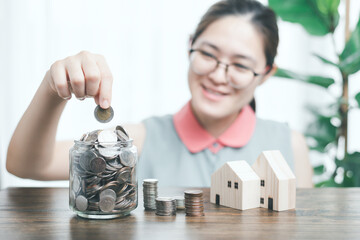 Asian woman smile and put coin in jar glass bank next to house model and stacked of coins on wood table and tree house decoration. Concept for loan, financial, real estate investment and money saving.