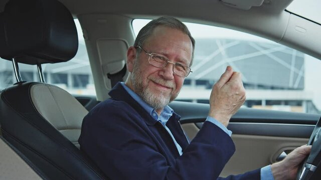 Portrait of tricky senior man sits in the luxurious modern car and making cash gesture asking money. Funny adult businessman. Gesturing. Wealthy lifestyle.