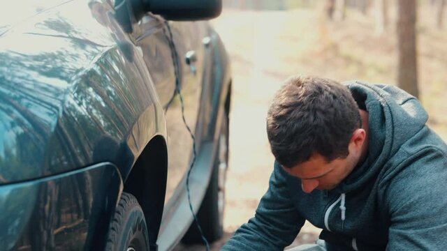 Young man and woman stand in forest. Slow motion of guy inflate car tires with air using compressor. Girl sitting inside car and showing they are getting late. Irritated man continue repairing.