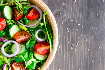 Fresh healthy salad with delicious ruccola, spinach, cabbage, arugula, feta cheese, red onion, cucumber, sesame seeds and cherry tomato on wooden background. Healthy and diet food concept.
