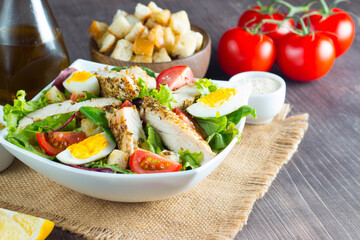 Fresh Caesar salad with delicious chicken breast, ruccola, spinach, cabbage, arugula, egg, parmesan and cherry tomato on wooden background. Oil, salt and pepper. Healthy and diet food concept.