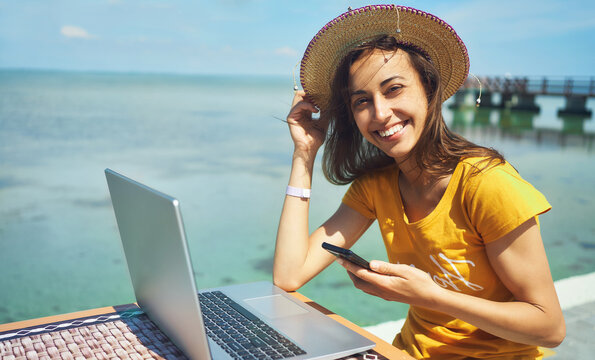 portrait happy freelancer woman in hat smiling to camera, holding smartphone, working on laptop computer by seashore. Freedom, remote job, freelance, travel and vacation concepts