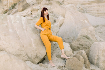 Summer bright portrait of beautiful dark haired girl in a yellow overalls posing in a sand quarry. Sexy woman in the desert leaning on the sand stone