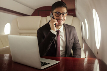 Handsome billionaire in his private jet, talking on phone with buisness partners, sitting at table with laptop ope