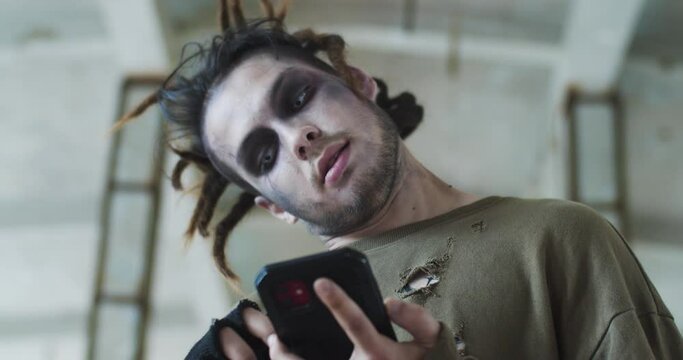 Terrifying evil zombie guy walking around the abandoned house. Pale infected living dead scrolling the news feed on the smartphone. Halloween, costume party, supernatural concept.