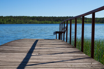 Obraz premium A wooden bridge with a railing and a bench leading into a blue lake with a view of the forest and the blue sky.