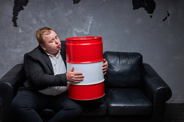 fat caucasian businessman hopes for a recovery in the oil industry, sits hugging oil barrel on sofa