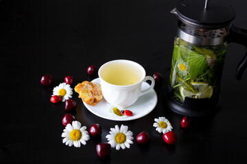 A white Cup with herbal tea, a dessert of toffee and corn sticks, and a chamomile with a Ladybird on a saucer, and a teapot with currant leaves, mint, chamomile and lemon.