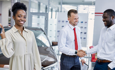 Fototapeta premium portrait of young black lady with keys of new car in hands, she looks at camera and smile. two men shake hands in the background. in cars showroom