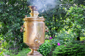 A traditional Russian samovar is heated by wood in the garden. A yellow retro samovar boils water...