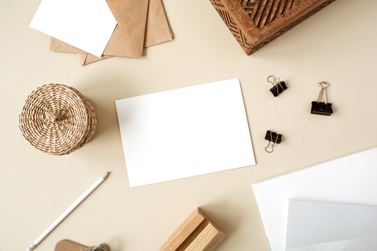 Blank Paper Sheet On Beige Table. Artist Home Office Desk Workspace With Wooden Casket, Pencil, Envelopes And Stationery. Webbing Invitation Card. Flat Lay, Top View Mockup With Empty Copy Space