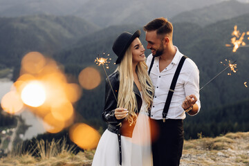 Beautiful bride and groom with sparklers on the top of the mountain. Wedding photoshoot. Smiling groom and bride. Blonde bride in black jacket .