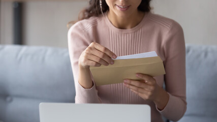 Crop close up of young Caucasian woman hold paper envelope open read postal mail letter at home,...
