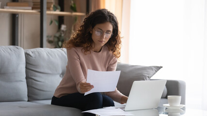 Focused young Caucasian woman in glasses sit on couch at desk work on laptop read paperwork,...