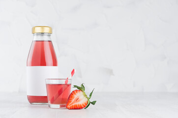 Fresh pink strawberry juice in glass bottle with blank label mock up with glass, straw, fruit slices in soft light white interior on wood table with copy space.