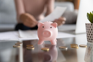 Crop close up of pink piggy bank on table with coins around, woman on background manage paperwork documents finances, female saving money, make investment in future, manage family budget