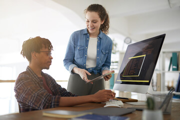 Portrait of smiling young woman talking to African-American man writing code while working in...