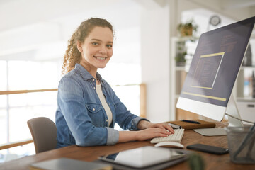 Warm-toned portrait of young woman using computer and smiling at camera while working at desk in...