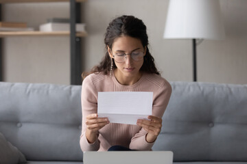 Serious young Caucasian woman in glasses sit on couch at home reading paper postal letter,...